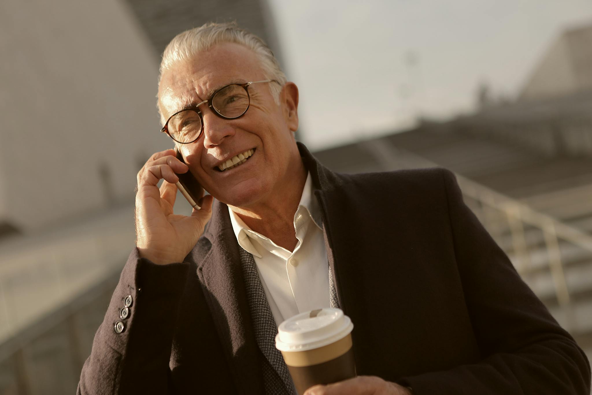 Elderly man smiling while talking on mobile phone with coffee in hand outdoors.